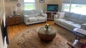 A living room with two white leather sofas, a glass coffee table with a floral centerpiece, a wicker side table, a flat-screen TV, and a large window with blinds. - FIFA Coming to America! Minutes to MetLife Stadium & NYC ACTION! LET’S GO! (Emerson)