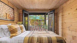 A wooden cabin bedroom with a bed, pillows, a blanket, a painting, and a balcony with a view of trees. - Charming Cabin Retreat • Pet-Friendly & Trails (Sevierville)