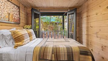 Une chambre dans une cabane en bois, avec un lit, des oreillers, une couverture, un tableau et un balcon donnant sur la forêt.