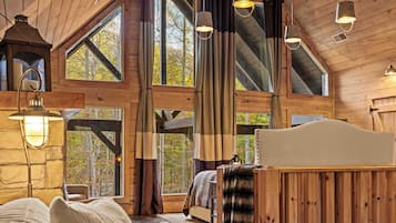 A cabin interior with a loft bed, large windows, wooden beams, and hanging light fixtures.