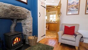 A cozy living room with a stone fireplace, a grey sofa with a red pillow, and a kitchen visible through an open door.