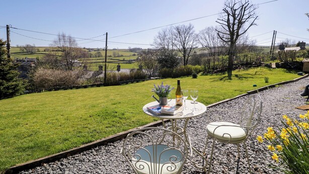 A patio with a table set for two, a bottle, and a book, overlooking a grassy area with a stone path and a view of the countryside.