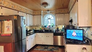 A kitchen with wooden cabinets, a stainless steel refrigerator, a sink, and a television displaying a program.