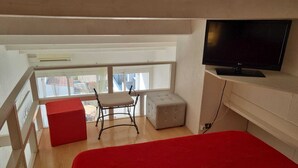 Een balkon met een rode bank, een witte poef, een zwarte stoel en een aan de muur bevestigde televisie.