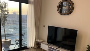 A modern living room with a large TV, comfortable seating, a dining area with a view of the ocean, and a large window with curtains.