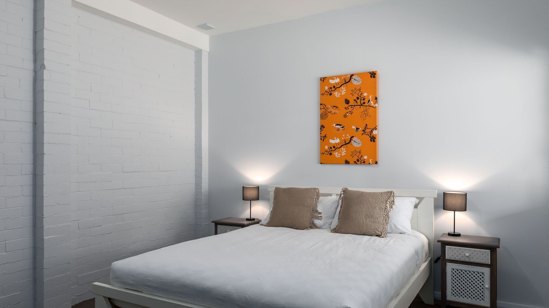 A bedroom with a white brick wall, a bed with white bedding, two nightstands with lamps, and an orange floral artwork on the wall.