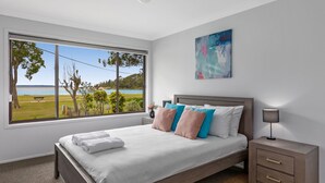 Un dormitorio con un ventanal grande, una cama con ropa de cama blanca, varios almohadones, una mesita de noche con una lámpara y un cuadro en la pared.