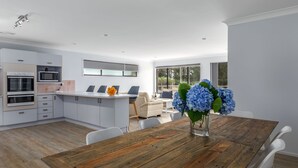 Cocina moderna con armarios blancos, horno empotrado y microondas. Hay un comedor con mesa y sillas de madera, y un jarrón con flores azules sobre la mesa.