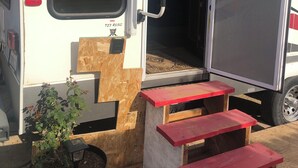 A white RV with a red and black stripe, a window with a view of palm trees, and a small wooden table with steps leading up to the door. - Cozy caravan with WiFi and AC in charming Thermal.Private location (Thermal)