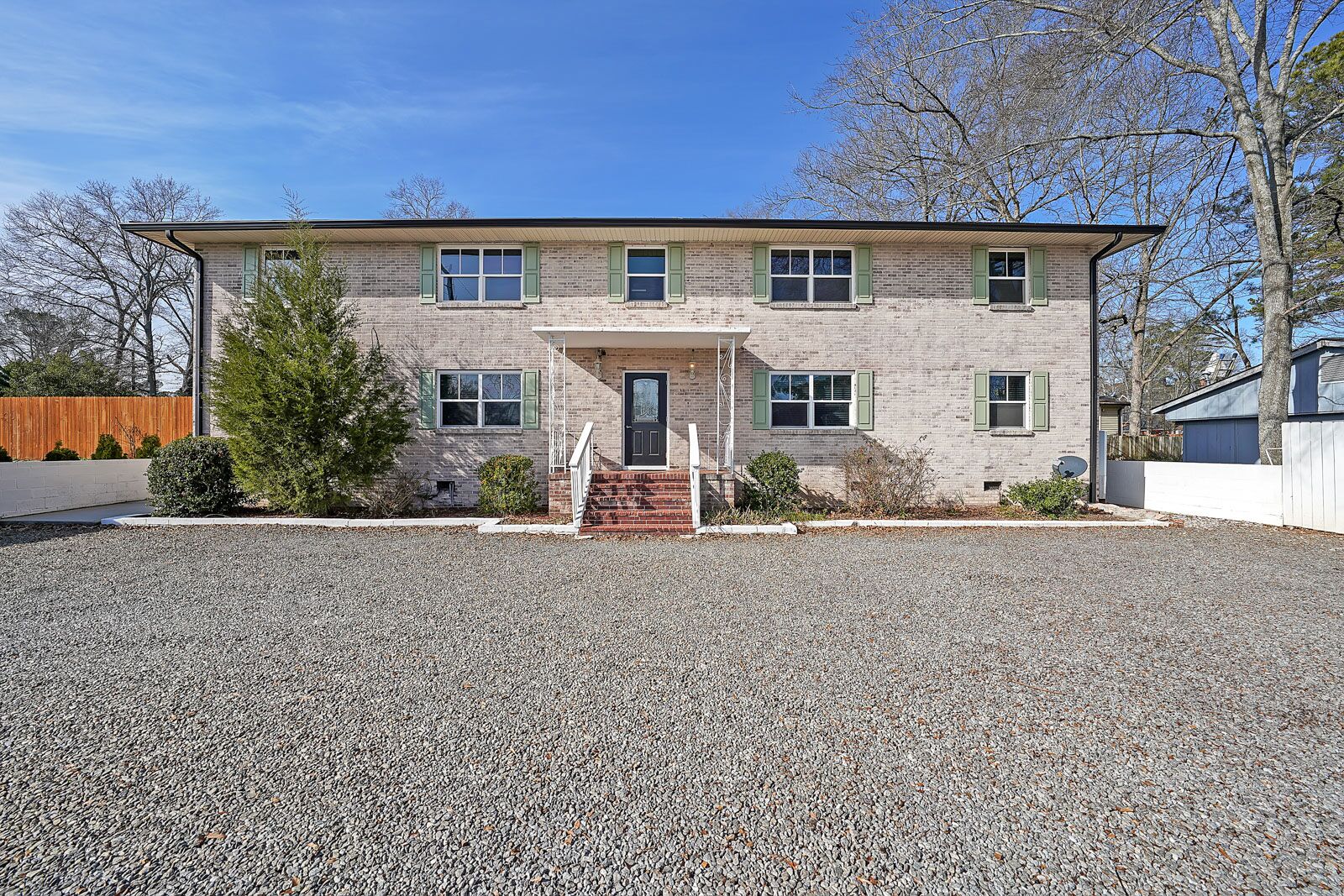 A two-story brick building with a front porch, a gravel driveway, and a wooden fence.