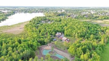 Luchtbeeld van een landelijk gebied met een groot waterlichaam, weelderige begroeiing en enkele gebouwen, waaronder een huis met een rood dak en zonnepanelen.