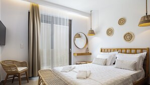A hotel room with a large bed, a wooden headboard, a bedside table with a lamp, a wicker chair, a bench, and a large window with curtains. - Feli Living - luxury villa Chania Crete Greece (Spilia)