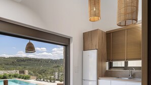 A modern kitchen with a large window, wooden cabinets, and a view of a swimming pool and landscape. - Feli Living - luxury villa Chania Crete Greece (Spilia)