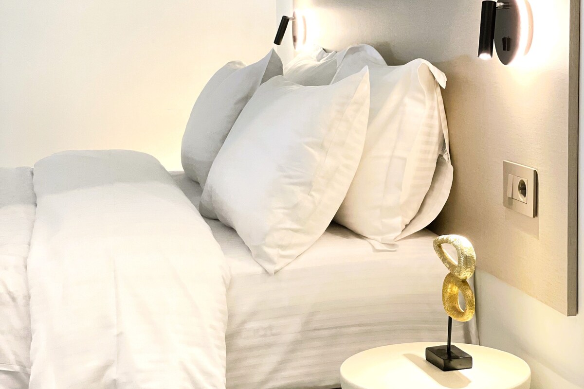 Un dormitorio moderno con cama, ropa de cama blanca, una mesita de noche con lámpara y un aplique de pared.