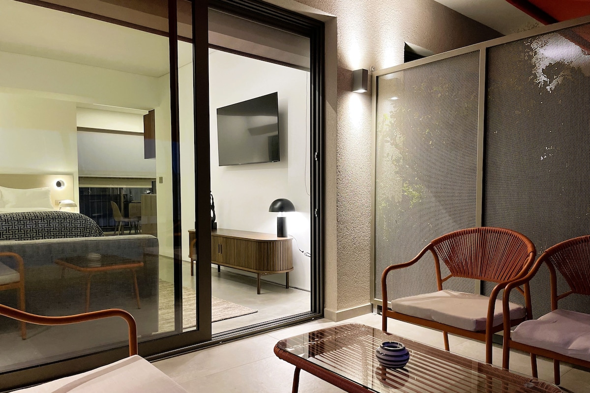 Ein modernes Hotelzimmer mit Glastür zu einem Balkon, einem Bett, einem Schreibtisch und zwei Korbstühlen.