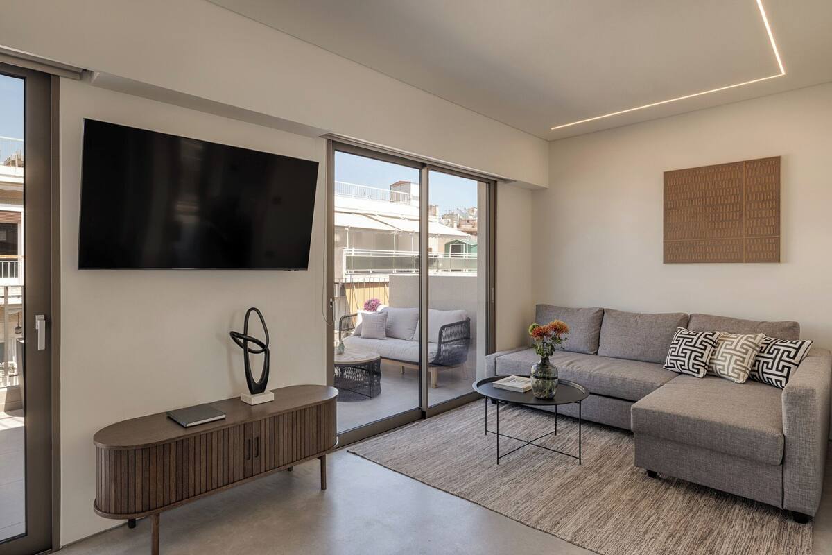 Uma sala de estar moderna com uma TV de tela plana, um sofá cinza, uma mesa de centro e uma porta de vidro deslizante que dá para uma varanda externa.