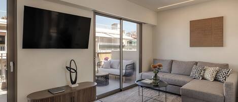 A modern living room with a flat-screen TV, a grey sofa, a coffee table, and a sliding glass door leading to an outdoor balcony.