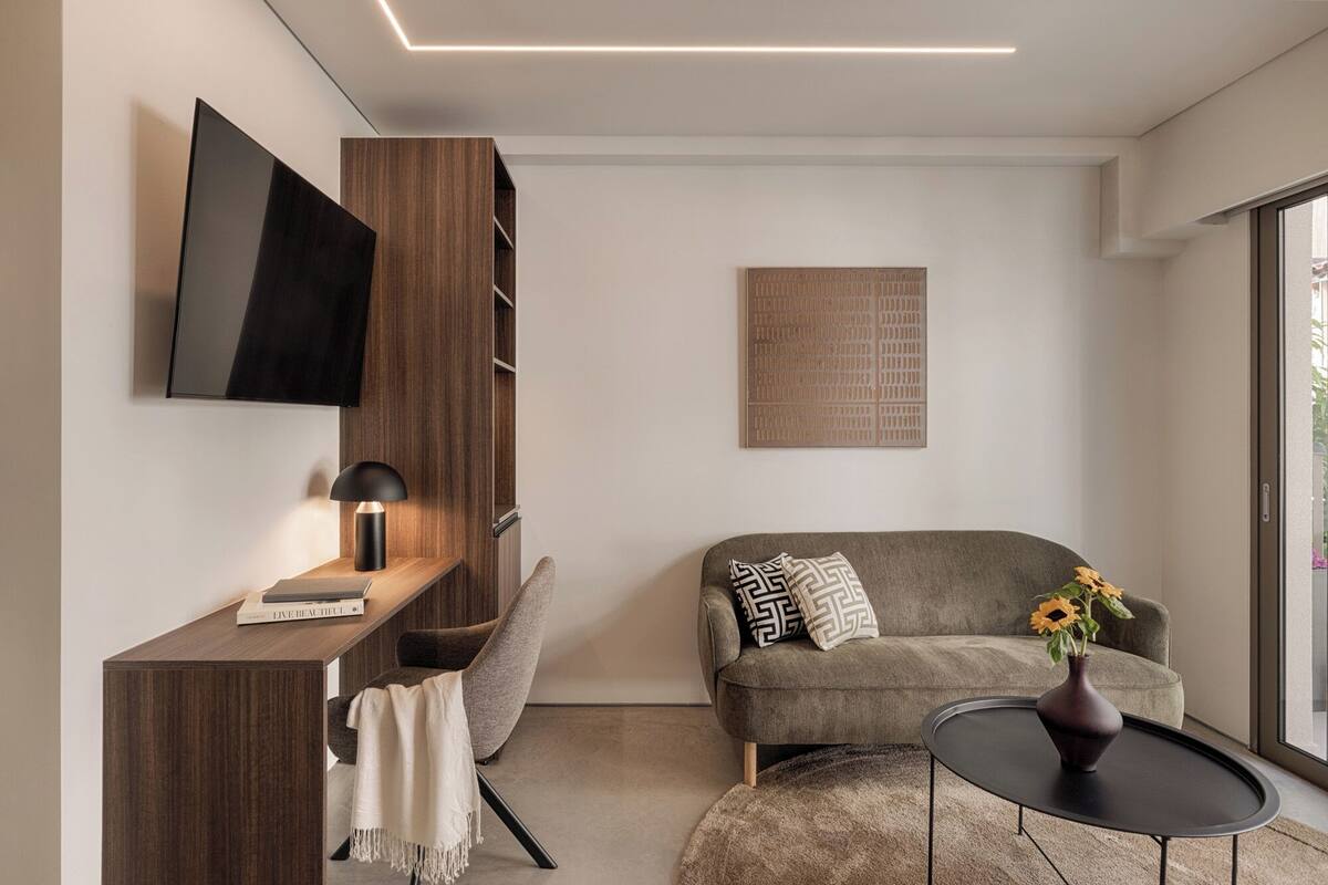 Uma sala de estar moderna com televisão de tela plana, uma estante, um sofá com almofadas estampadas, uma mesa redonda pequena com um vaso, e uma janela grande.