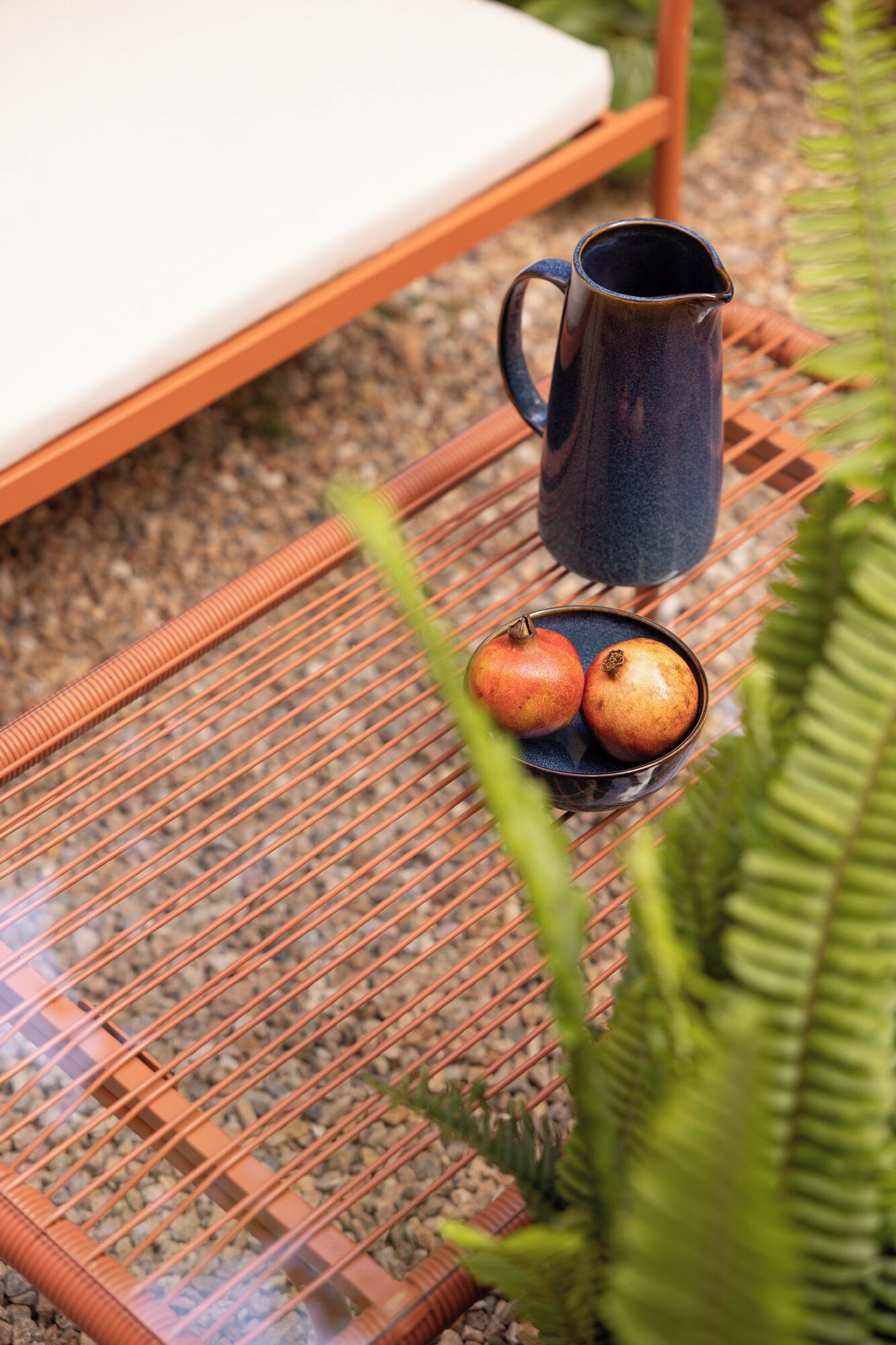 A wicker table with a dark ceramic pitcher and a bowl containing two pomegranates, set on a patterned mat.