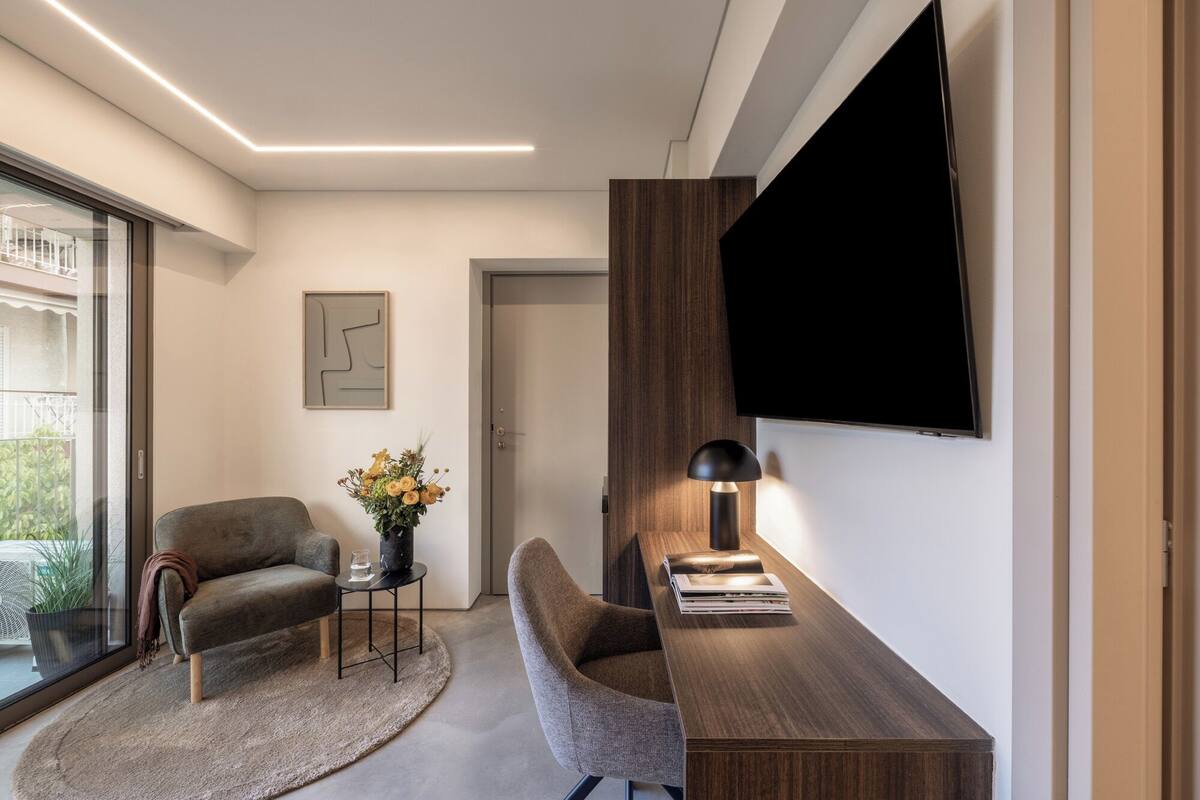 Quarto moderno com escrivaninha, duas cadeiras, televisão, um vaso de flores e um relógio na parede.