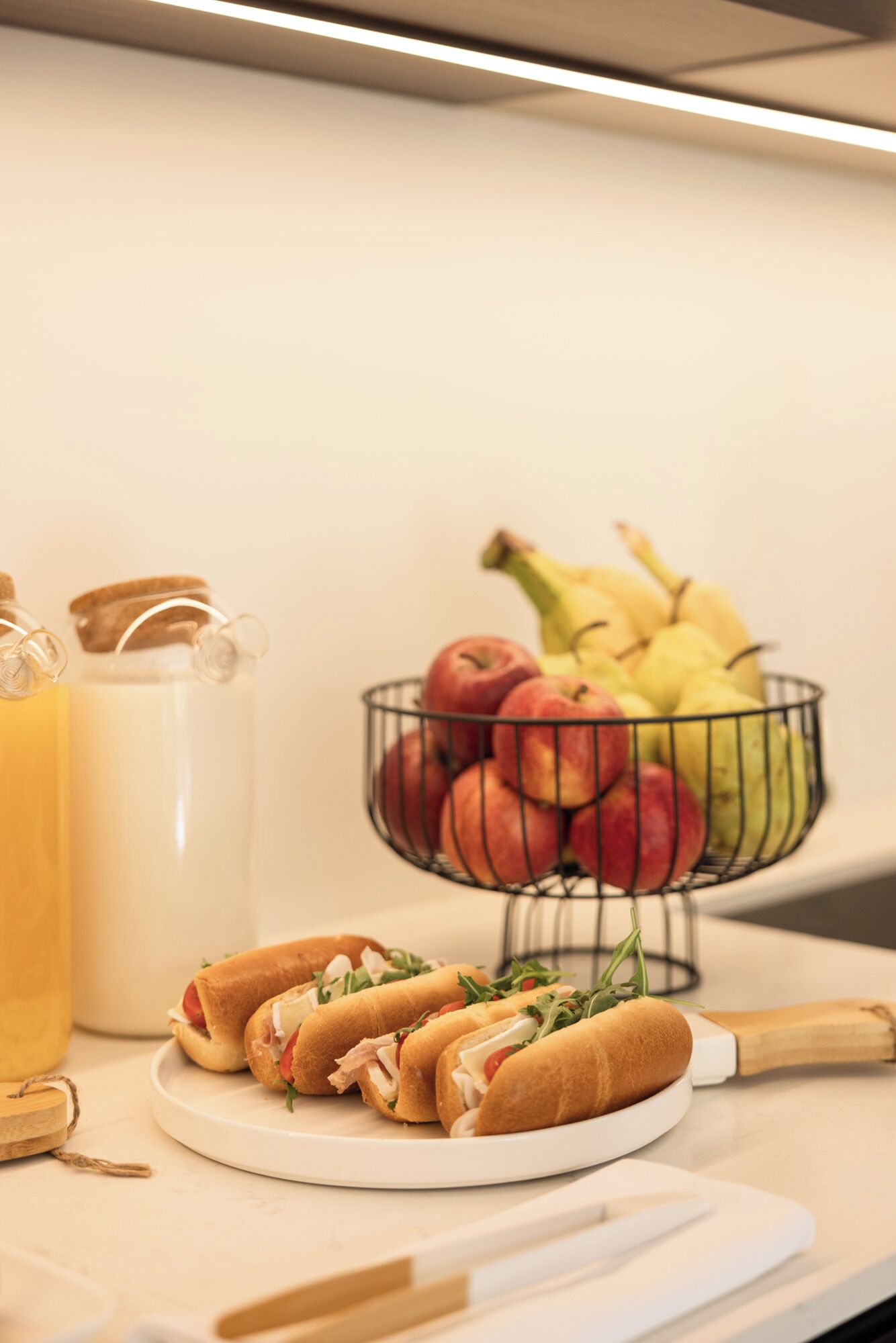 A kitchen counter with a plate of sandwiches, a fruit bowl, and a pitcher of juice.