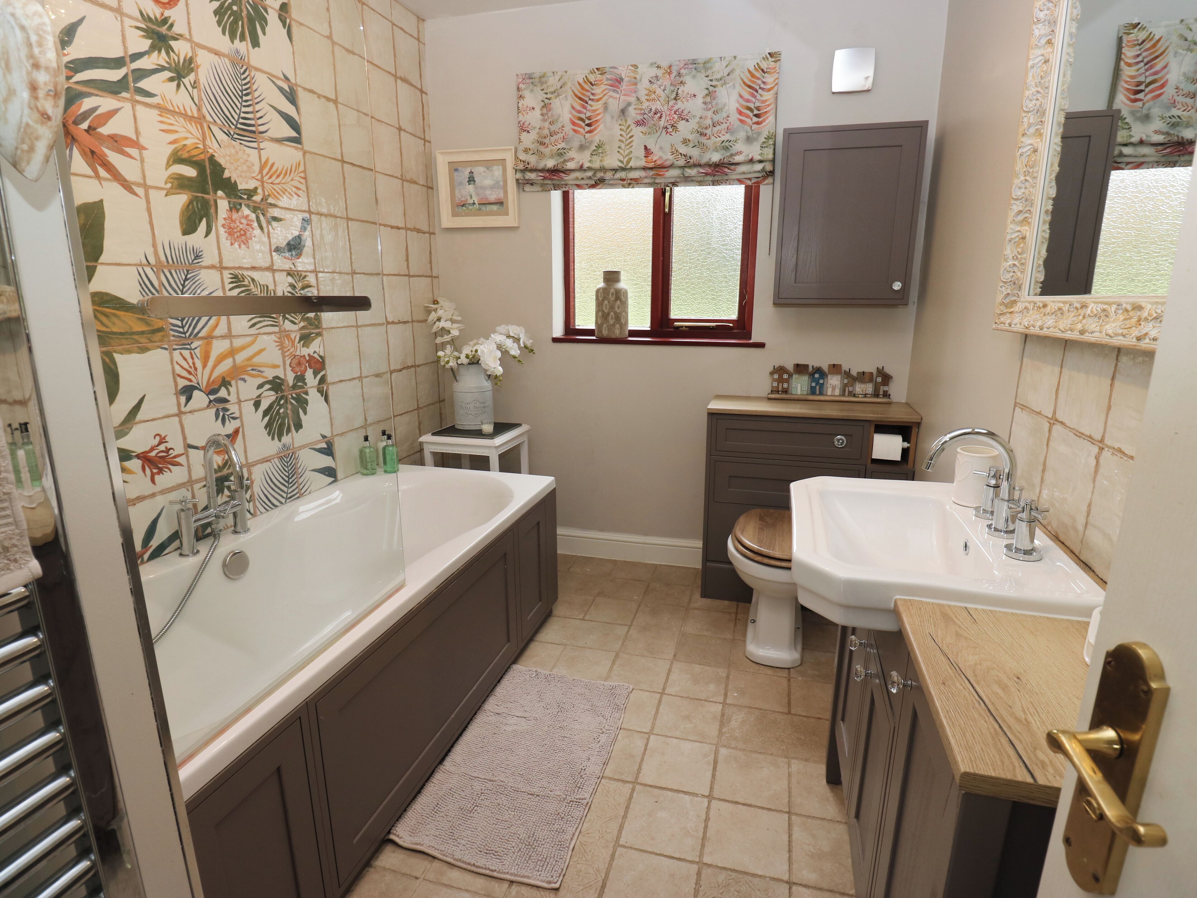 A bathroom with a bathtub, toilet, and sink, featuring floral tiles, a window with a view, and a small shelf with decorative items.