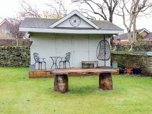 A rustic outdoor seating area with a wooden table and metal chairs, a hanging chair, and a clock on the wall.