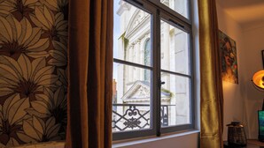 Ein Zimmer mit Fensterblick auf ein Gebäude, Vorhängen, einem kleinen Tisch mit Lampe und einer Wand mit floralem Muster.
