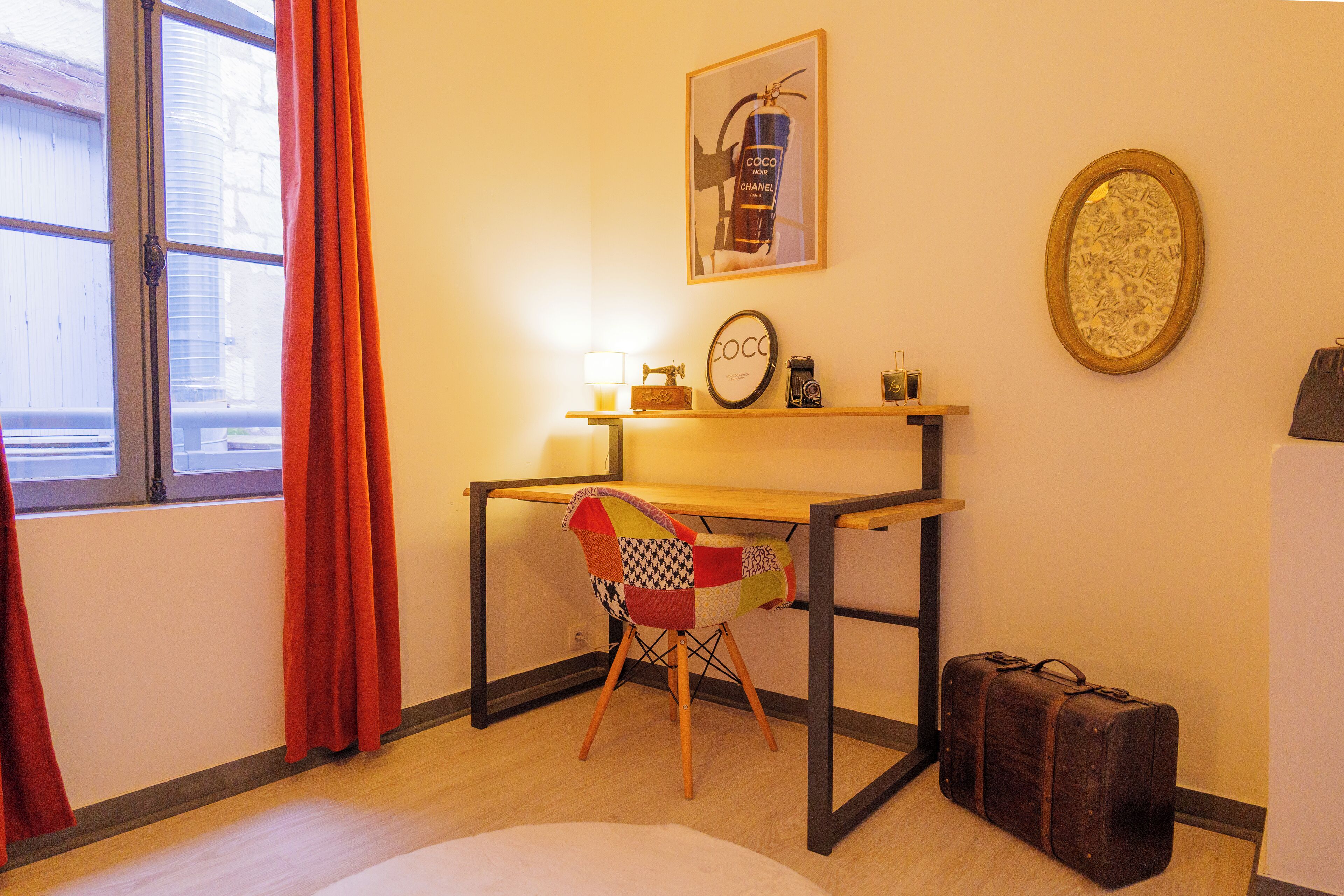 A room with a wooden desk, a chair with a patchwork seat, a suitcase, a framed picture of a Chanel bottle, and a window with curtains.