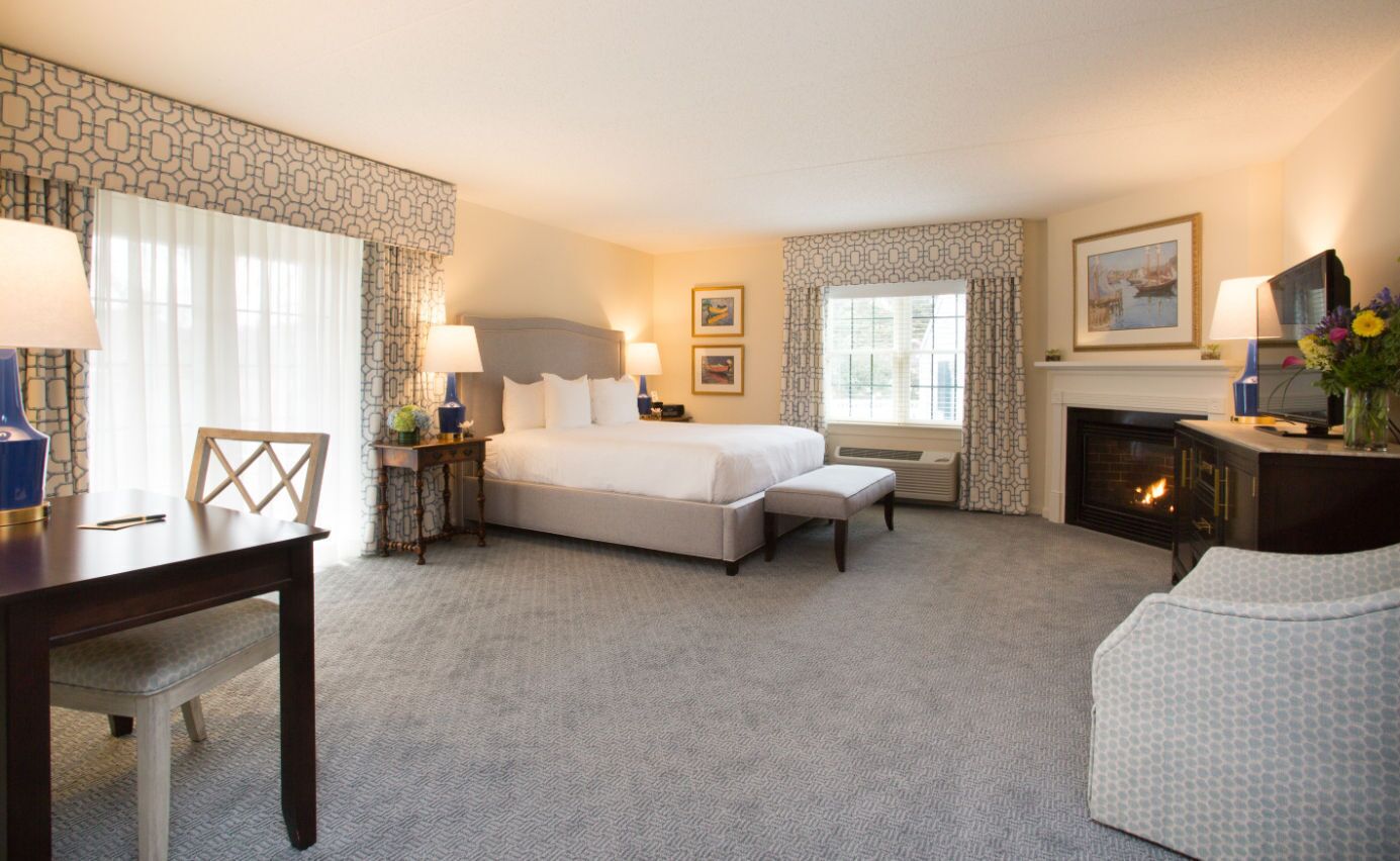 A hotel room with a large bed, a desk, a chair, a fireplace, and a window with curtains.