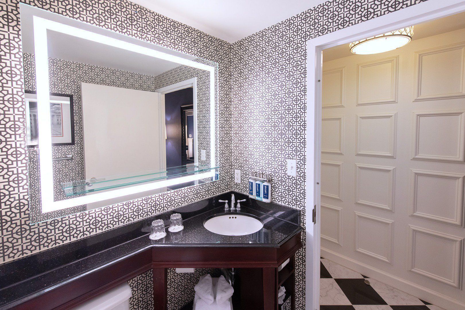 Un baño con pared de diseño blanco y negro, un espejo grande, encimera negra y lavamanos blanco.
