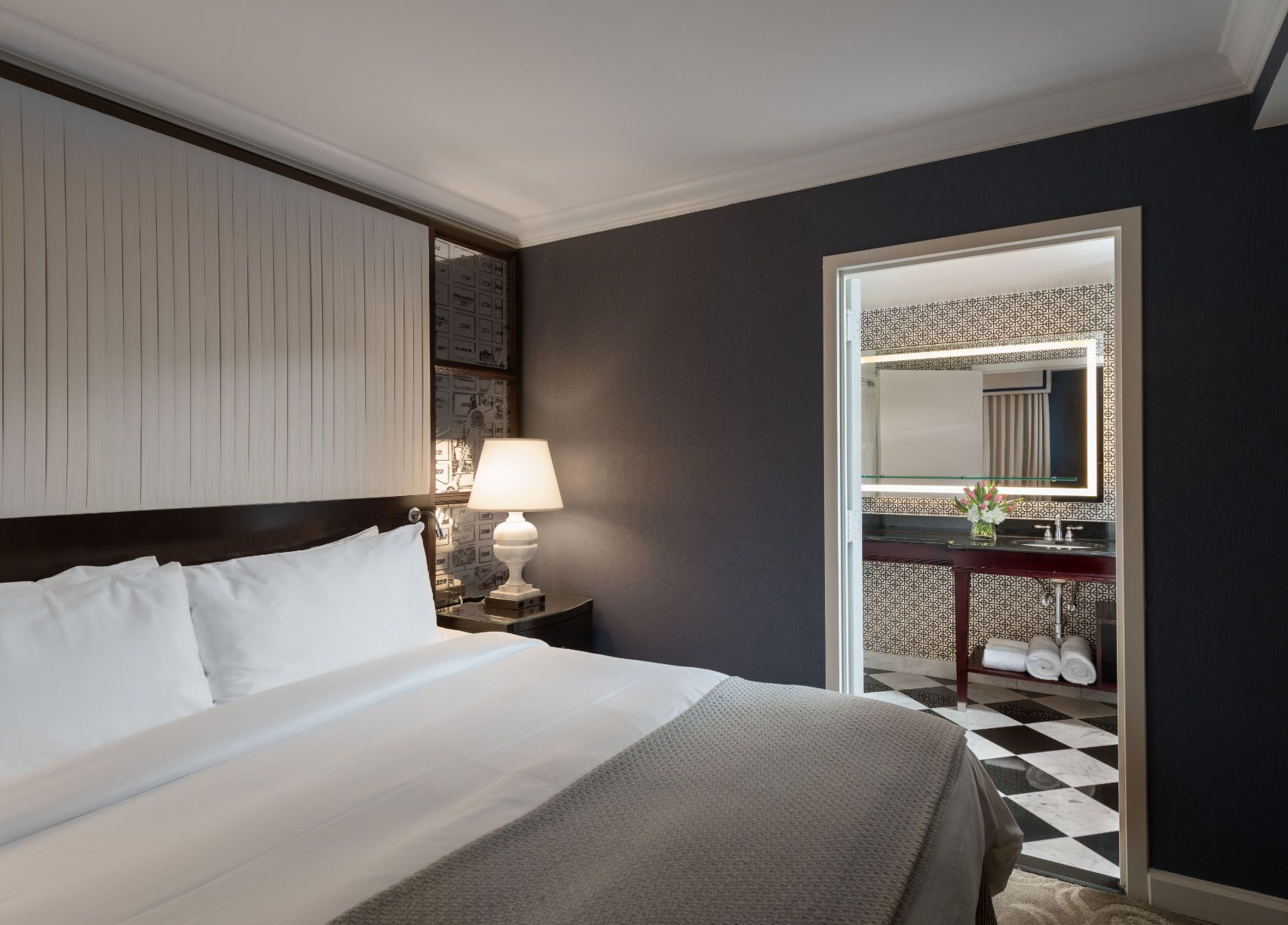 A hotel room with a bed, a nightstand with a lamp, a bathroom with a mirror, and a checkered floor.