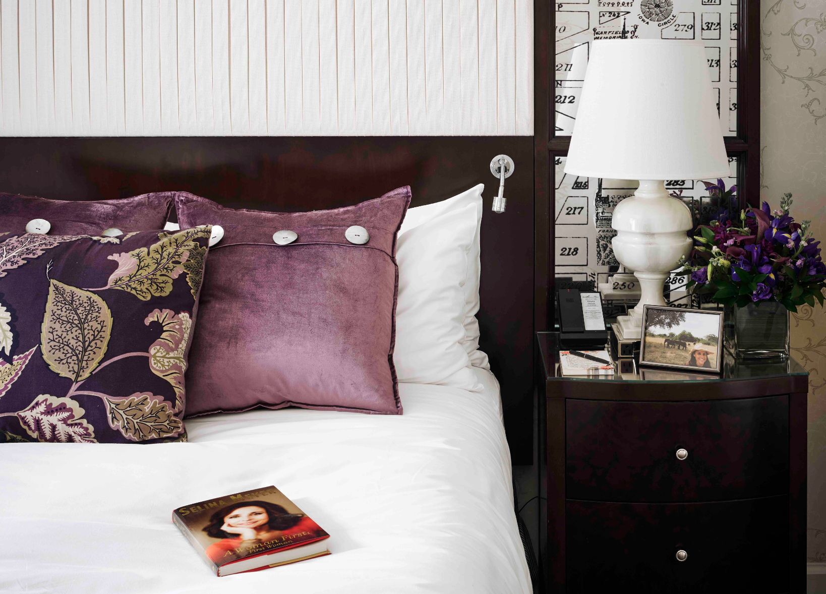 A neatly made bed with a purple floral pillow and a book titled "A Woman's First".