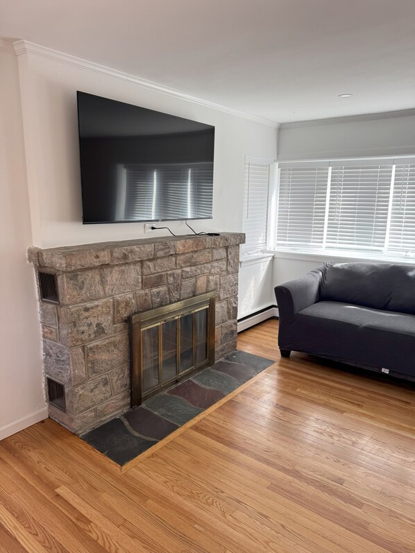 A living room with a stone fireplace, a flat-screen TV mounted above it, a dark-colored sofa, and a window with blinds. - Spacious NYC Getaway House (Yonkers)