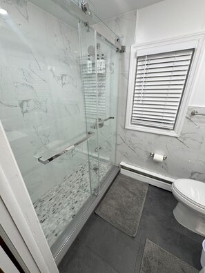 A modern bathroom with a glass-enclosed shower, a toilet, and a window with blinds. - Spacious NYC Getaway House (Yonkers)