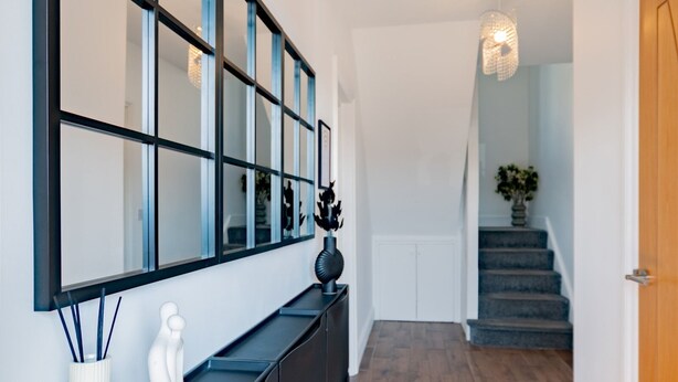 Un couloir moderne avec un miroir à cadre noir, un vase blanc et une sculpture noire posée sur une console sombre. Un escalier recouvert d’une moquette bleue mène à un palier où se trouve une plante en pot.