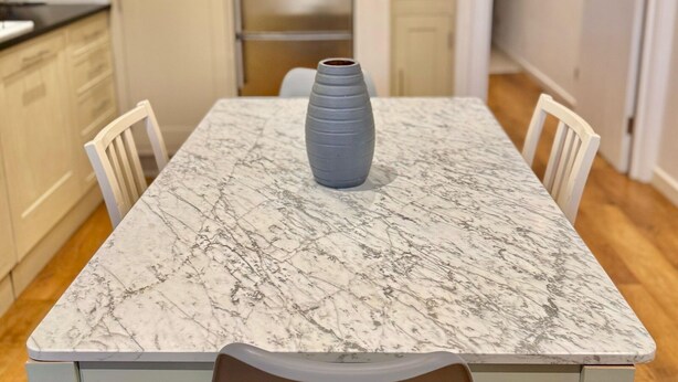 Een eettafel met een marmeren blad, omringd door witte stoelen, in een keuken met lichtgekleurde kastjes en een roestvrijstalen koelkast.