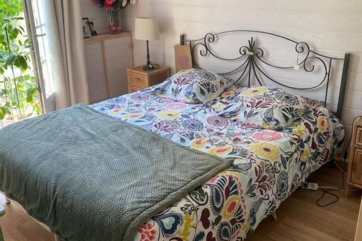 Een slaapkamer met een bed, een nachtkastje, een lamp, een rieten stoel en een raam met gordijnen.