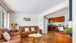 A living room with a brown leather sofa, a round coffee table, and a kitchen area with wooden cabinets and a white refrigerator.