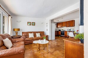 A living room with a brown leather sofa, a round coffee table, and a kitchen area with wooden cabinets and a white refrigerator.