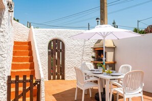 A patio with a table and chairs, a barbecue grill, and a white wall with a textured surface. - Villa Bourgon by Villa Plus (Cala en Porter)