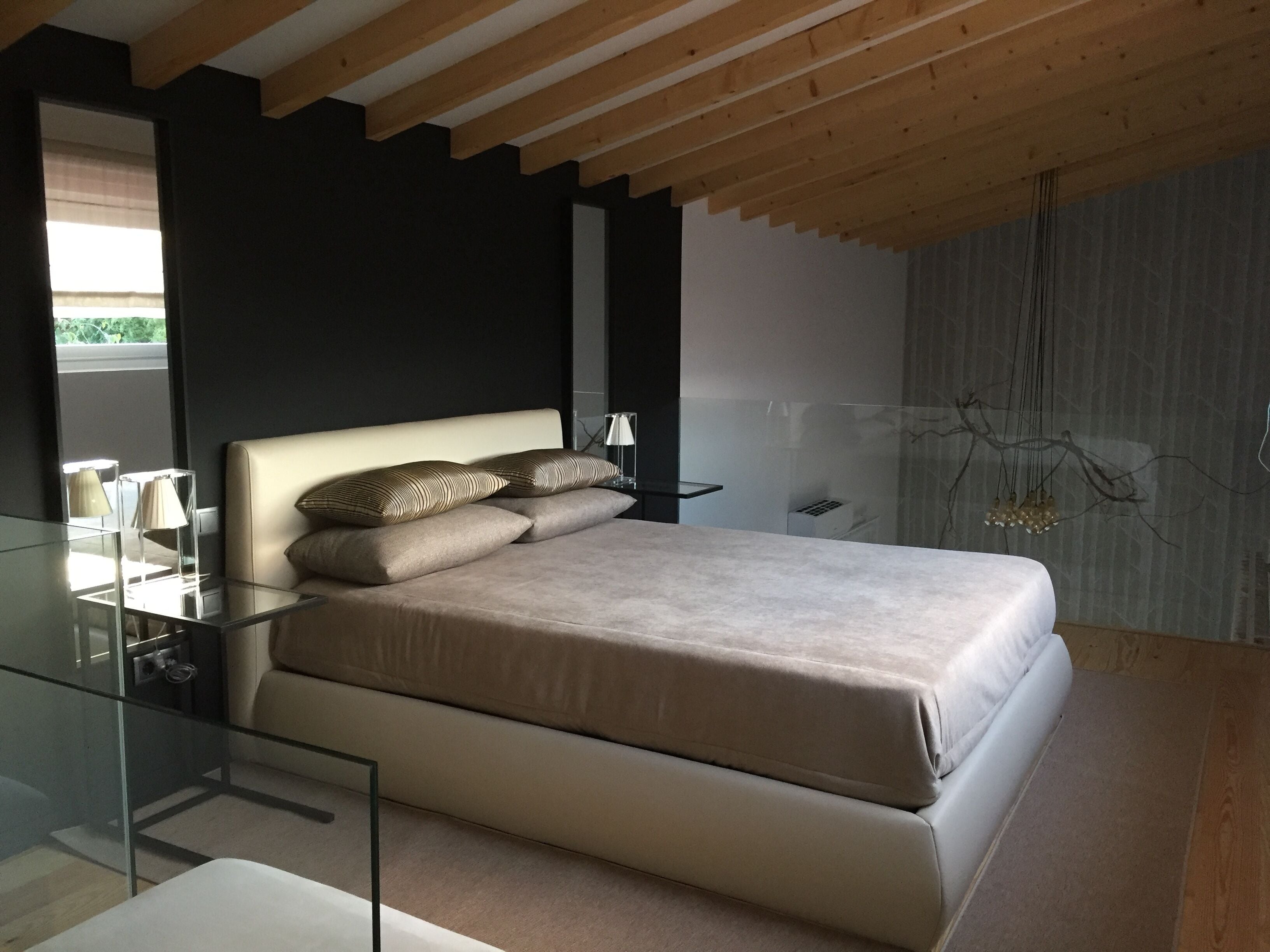 A modern bedroom with a glass bedside table, a bed with a beige headboard, and a dark wall.