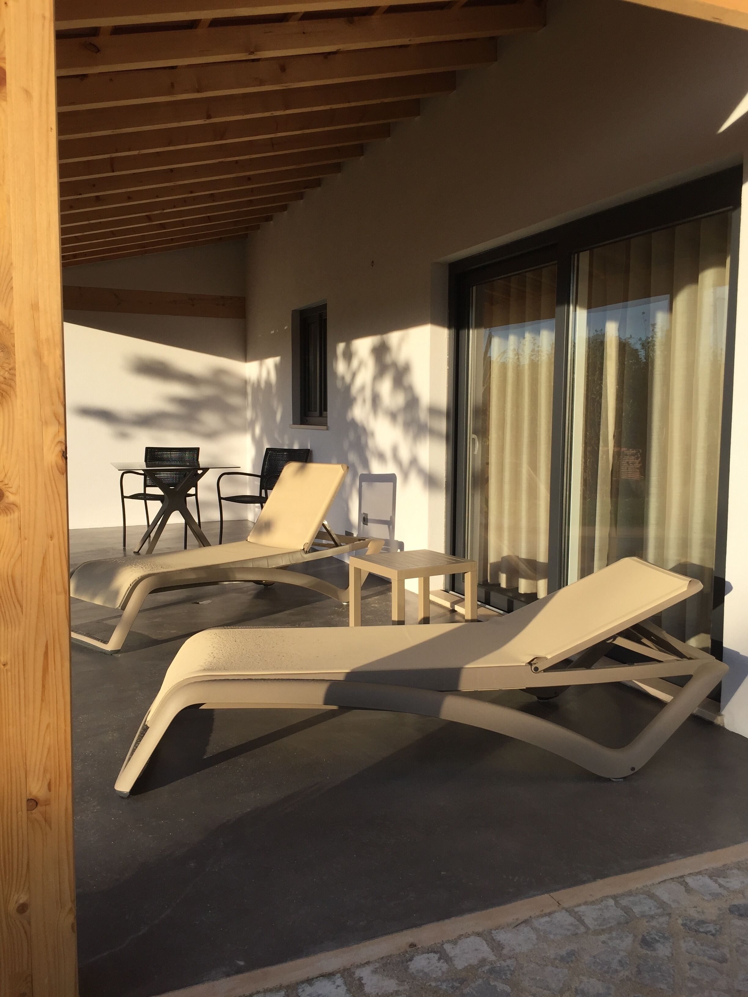 Un espacio de estar techado al aire libre con sillones reclinables y una mesa, una ventana y una puerta con cortinas.