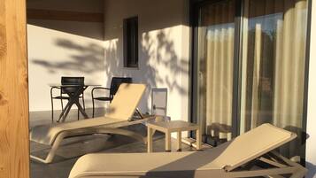 Un espacio de estar techado al aire libre con sillones reclinables y una mesa, una ventana y una puerta con cortinas.