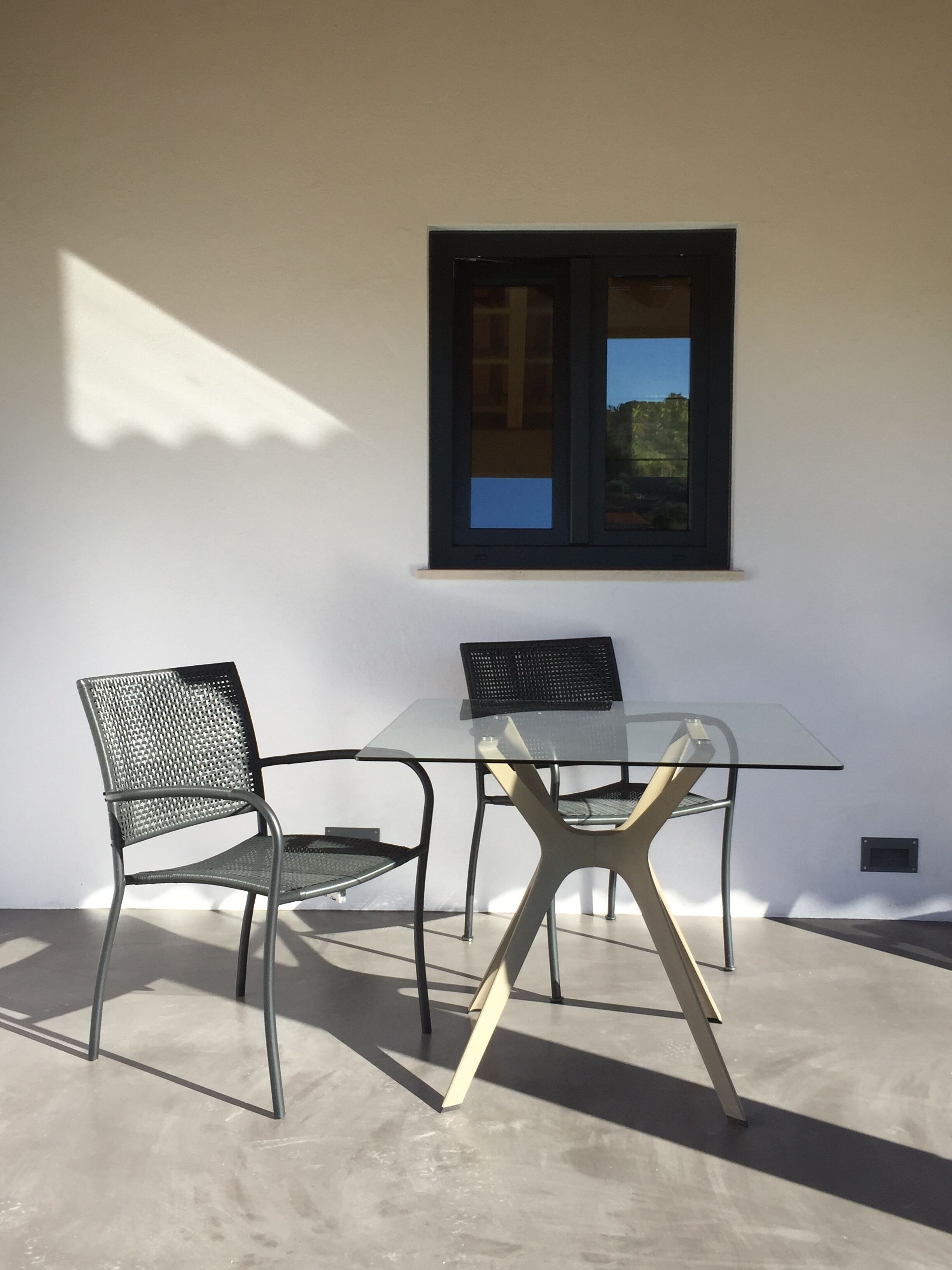 A modern outdoor seating area with a glass table and metal chairs, a window with a view, and a concrete floor.