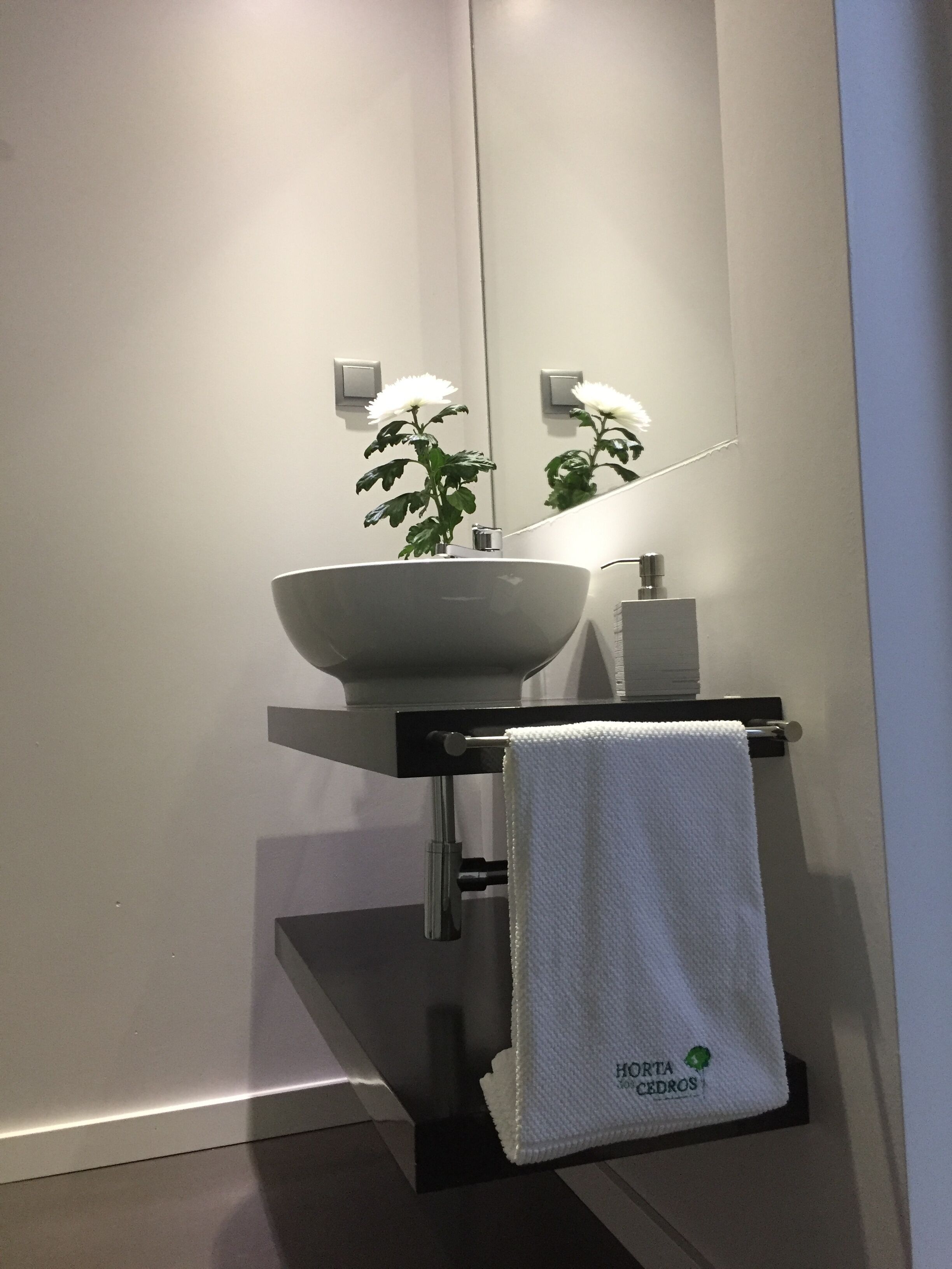 Un baño moderno con un lavamanos blanco, un perchero con una toalla blanca y un espejo que refleja una flor blanca.