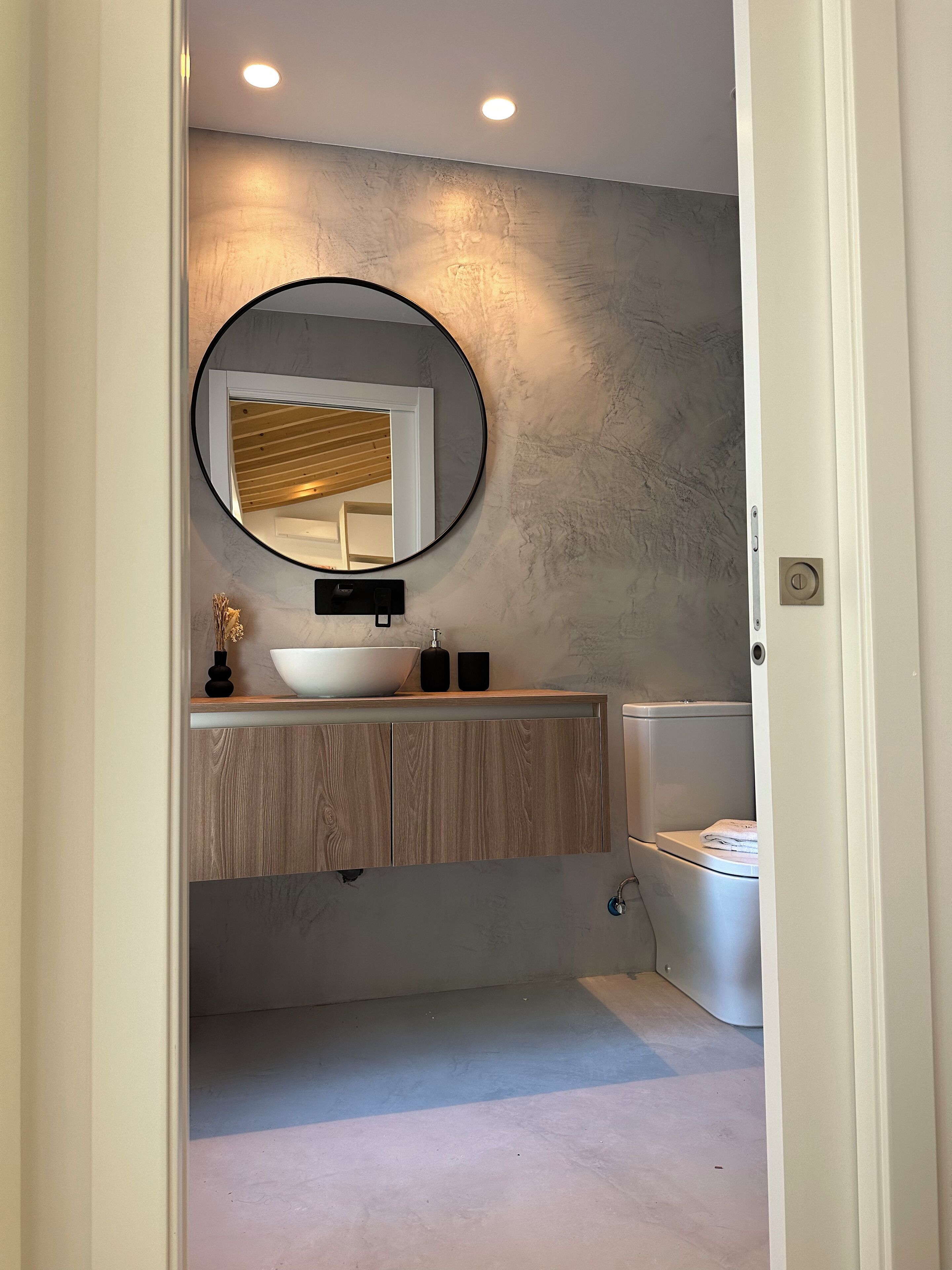 Un baño con espejo redondo, tocador de madera, lavamanos blanco y váter.