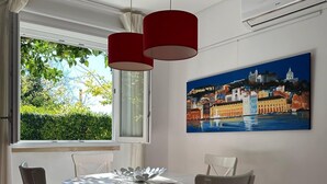 A dining area with a round table, white chairs with red cushions, a painting of a cityscape, and a window with a view of greenery. - Horta dos Cedros  (Tomar)