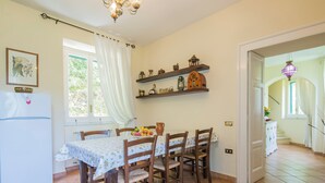 Zona pranzo con tavolo apparecchiato per quattro, circondato da sedie di legno. Un frigorifero bianco si trova alla sinistra e sopra il tavolo ci sono scaffali con diversi oggetti da cucina.