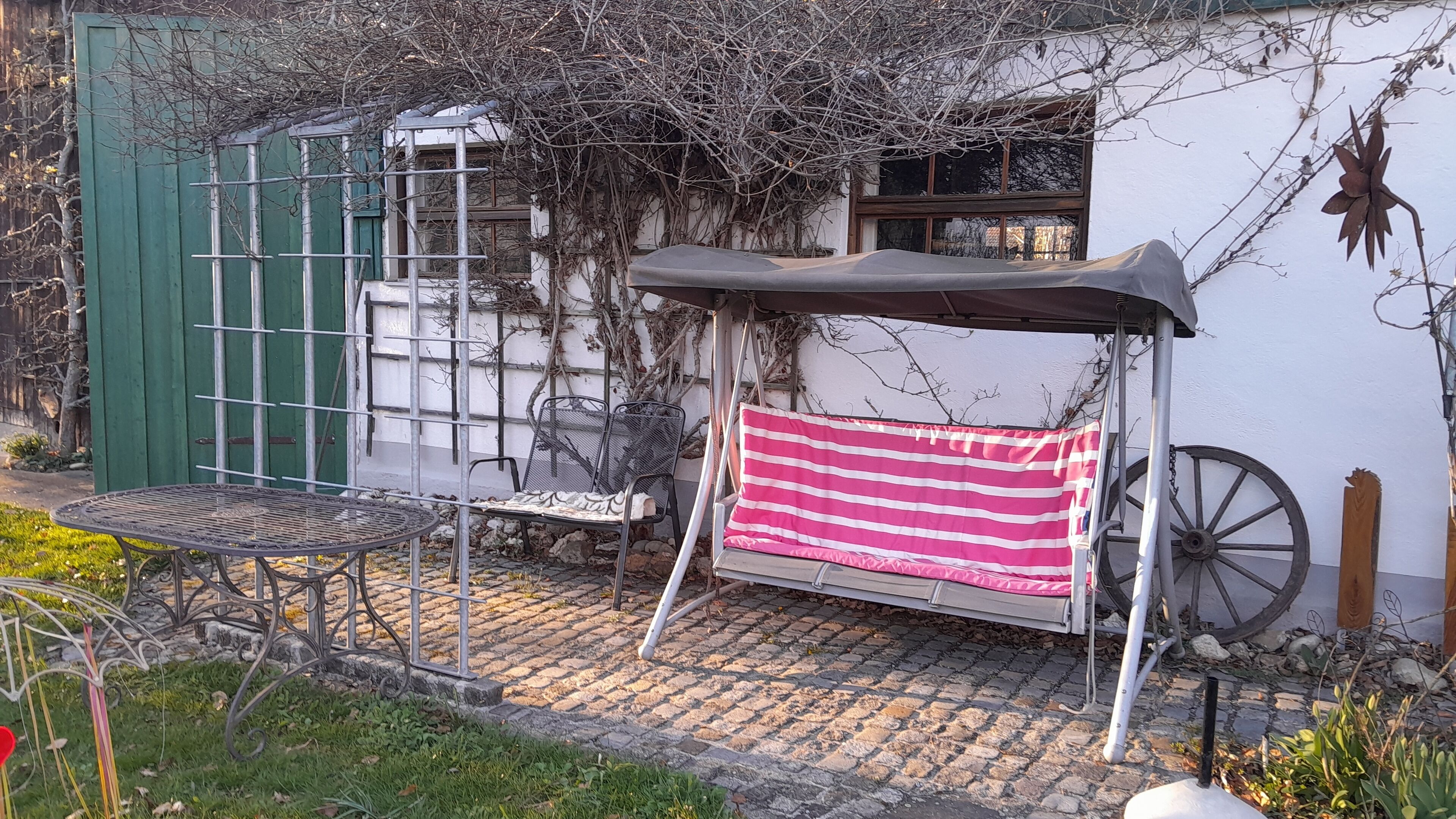 Un espacio de estar al aire libre cubierto con un cojín a rayas, una mesa metálica y sillas, rodeado por un jardín y un edificio con una puerta y ventanas verdes.